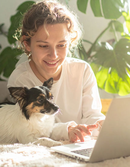 Femme souriante utilisant un ordinateur portable avec son petit chien à ses côtés, moment calme et complice à la maison.